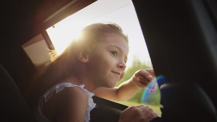 Little girl looks at scenic countryside views at sunset through open car window waving hello. Happy child enjoys going on road trip with family during summer vacation sitting on backseat closeup