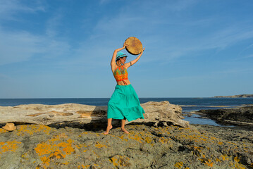 A woman in boho style clothes with a shamanic ritual tambourine on the rocks on the seashore on a clear sunny day. The concept of freedom of mind and body in nature