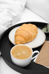 Tray with tasty croissant, cup of coffee and envelope on white bed