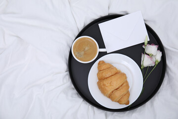 Tray with tasty croissant, cup of coffee and flowers on white bed, top view. Space for text