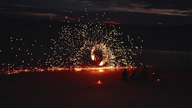 A street performer dances with fire at night on the ocean beach. Sparks of fire fly around the man, a mesmerizing sight.