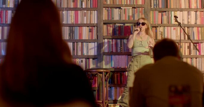Woman stand-up comedian glasses on eyes on microphone monologue stage. She speaks, gesticulates. The reaction of the audience in bokeh. Dark room.