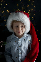 A cute boy in a Santa Claus hat smiles slyly against the backdrop of a Christmas tree and lights