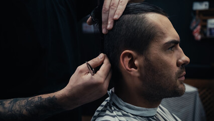 Hairstylist holding scissors and combing hair of man in hairdressing cape in salon.