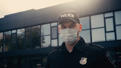 police officer in medical mask and cap with lettering looking at camera near building.