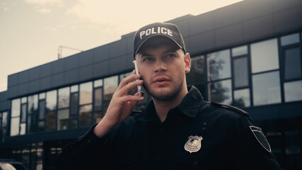 serious policeman in uniform and badge talking on smartphone on urban street.