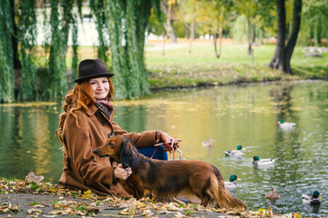 Young red-haired woman sitting near a small river in the park with a red dachshund dog. 