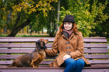 Young red-haired woman sit on a bench in autumn park with a red dachshund dog. 