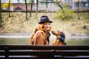 Young red-haired smiling  woman sit on a bench in autumn in the park with a red dachshund dog. 