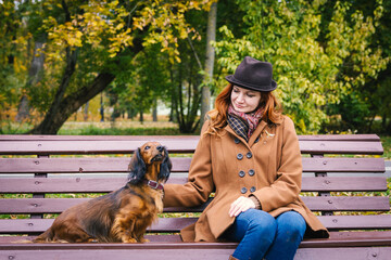 Young red-haired woman sit on a bench in autumn park with a red dachshund dog. Woman looks at a pet.