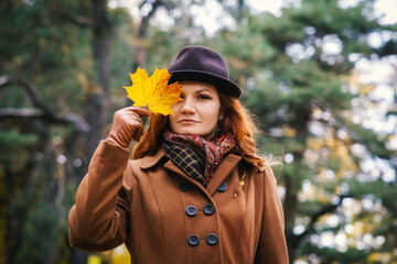 Portrait of a young red-haired woman in a hat against the background of green pine-trees. Woman posing covering one eye with a maple leaf
