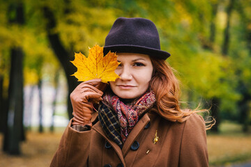 Portrait of a young red-haired woman in a hat against the background of autumn foliage. Woman posing covering one eye with a maple leaf
