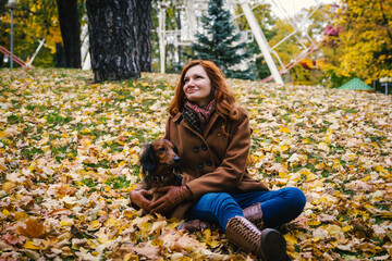 Young red-haired woman with a red dachshund dog sit on a grass in autumn in the park. 