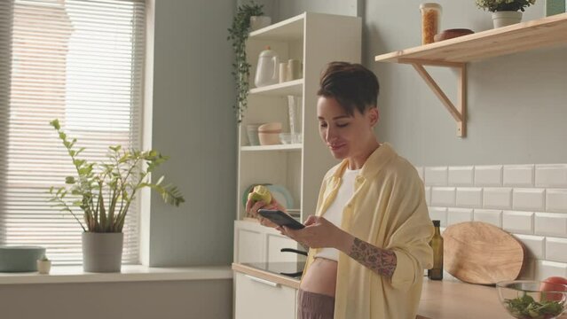 Medium shot of cheerful young pregnant woman eating apple and texting with friends on smartphone standing at minimalist kitchen