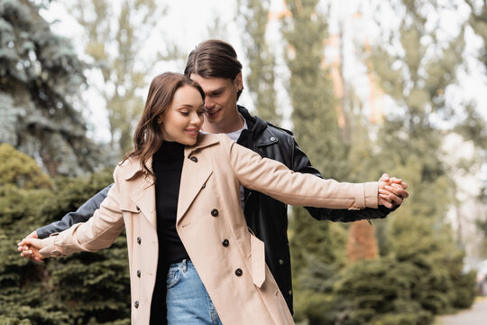 cheerful man holding hands with pleased girlfriend in beige trench coat.
