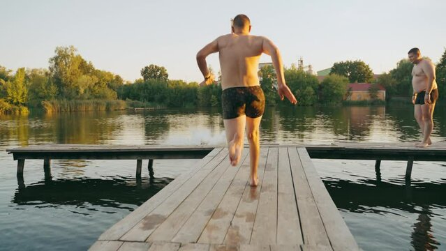 Young guys jump into the water from a wooden pier doing tricks.