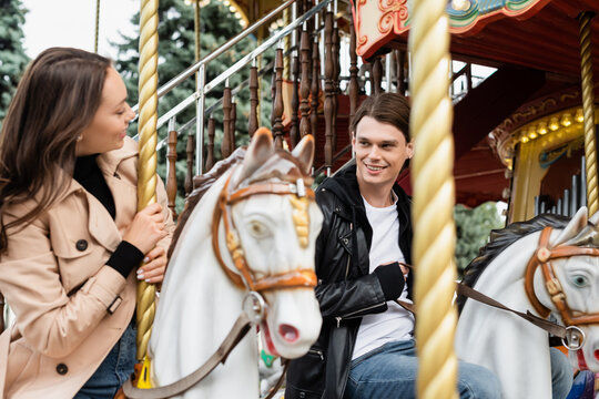 cheerful young man looking at girlfriend riding carousel horse in amusement park.