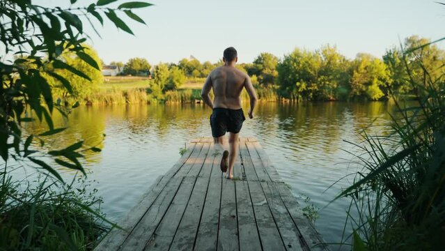 Back view of a young guy jumping from a wooden pier into a lake.