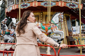 side view of curly young woman in beige trench coat standing near carousel in amusement park.