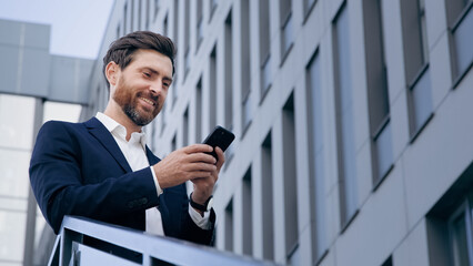 Stylish handsome businessman holding phone reading email stands on street smiling writes message...