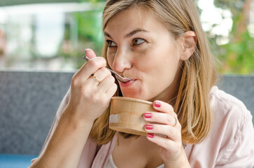 Cheerful woman with blond hair and red nails eats strawberry ice cream with a spoon in a cafe