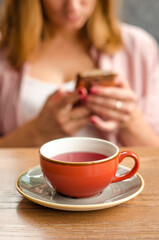 Obraz na płótnie Canvas Red cup of red tea, a gray teapot stand on a table in a cafe. In the background, a young woman is working on her smartphone