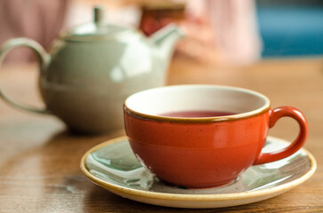 Red cup of red tea, a gray teapot stand on a table in a cafe. In the background, a young woman is working on her smartphone