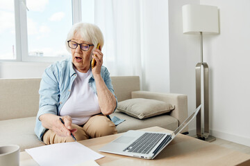 an elderly woman engaged in a telephone conversation is actively gesticulating with her hand while sitting on a cozy sofa in a bright interior with a laptop on the table