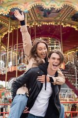 excited man in black jacket piggybacking happy girlfriend in amusement park.