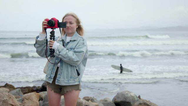 Shot of a young blonde woman shooting filming herself by camera on the ocean beach. Big Sur California coast. Waves crashing. Travel blogger on vacation. High quality 4k slow motion footage.