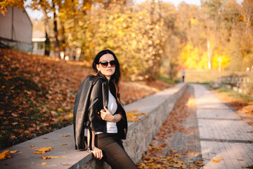 Fototapeta na wymiar Portrait of young stylish woman wearing sunglasses and leather jacket standing in park during sunny weather in autumn