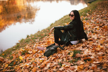 Young thoughtful stylish woman looking away while sitting on fallen leaves by the lake in park during sunny weather in autumn