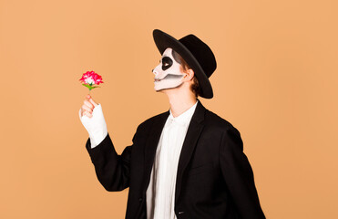 Portrait of spooky handsome guy made makeup for Halloween event, has image of ghost, red rose flower in hand, wears black hat, has scary look, dressed in zombie attire