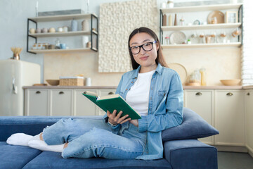 Young beautiful Asian woman in denim clothes and glasses sitting on sofa at home. He holds a green book in his hands, looks at the camera, smiles. Rest for the weekend.