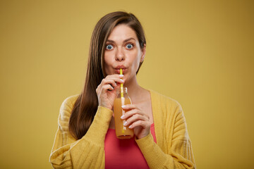 Surprising woman drinking orange juice from glass bottle with straw. Eyes wide open. Isolated advertising portrait on yellow.