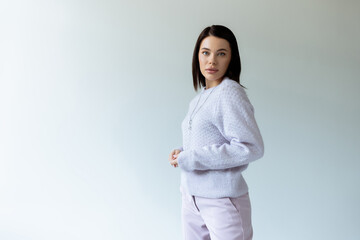 brunette woman in knitted sweater and trousers looking at camera on grey background.