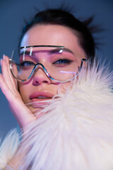 portrait of young woman in transparent sunglasses and white faux fur jacket looking at camera on blue background.