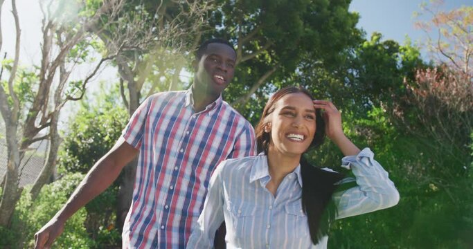 Video of happy diverse couple holding hands and walking in sunny garden smiling