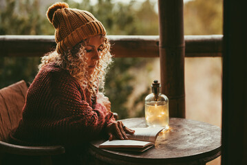 One woman reading a book outdoor sitting a little wooden table and wearing sweater and wool hat for cold temperature. Female adult people and education in leisure activity at home. Pretty lady alone