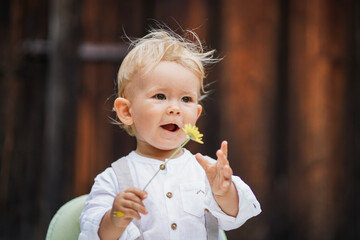 one year old happy blond german baby boy in white shirt playing outside infront of a rustic wooden hut gate with a yellow flower to congratulate for birthday