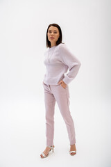 Fototapeta na wymiar full length of trendy brunette woman in knitted sweater and pants posing with hand in pocket on white background.