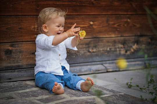 one year old happy blond baby boy in white shirt sitting outside on the ground infront of a rustic wooden hut and holding a yellow marguerite flower to send love and happiness