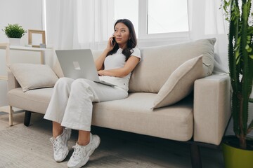 Fototapeta premium Young Asian woman looking at laptop screen on her lap sitting on couch at home lifestyle work as a freelancer