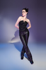 full length of glamour woman in leather pants and black corset posing with hands behind back on purple background.
