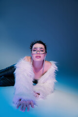 glamour woman in white faux fur jacket and transparent eyeglasses lying on blue background.