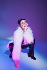 trendy woman in transparent eyeglasses and white fluffy jacket looking at camera on purple background.