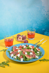Fototapeta na wymiar Autumn figs salad with arugula, feta, raw ham in blue plate on yellow and blue background served with rose wine. Flexitarian diet, paleo diet fall salad, copy space