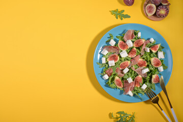 Autumn figs salad with arugula, feta, raw ham in blue plate on yellow background. Flexitarian diet, paleo diet fall salad, top view, flat lay with copy space