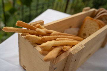 Bread sticks and bread served in wooden container on a white restaurant tablecloth. In the background the green of the surrounding nature. Focus on bread sticks.