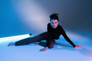 brunette barefoot woman in black sweater and pants sitting near fluorescent lamp on blue background.